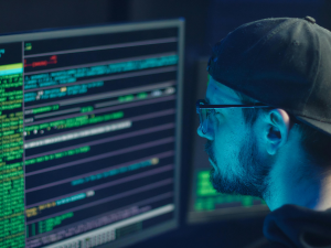 computer screen with code, man with black ball cap and glasses looking at screen